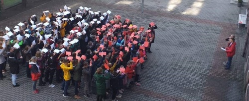 Uczniowie stoją na podwórzu szkolnym i trzymając w górze kartki tworzą flagę biało-czerwoną